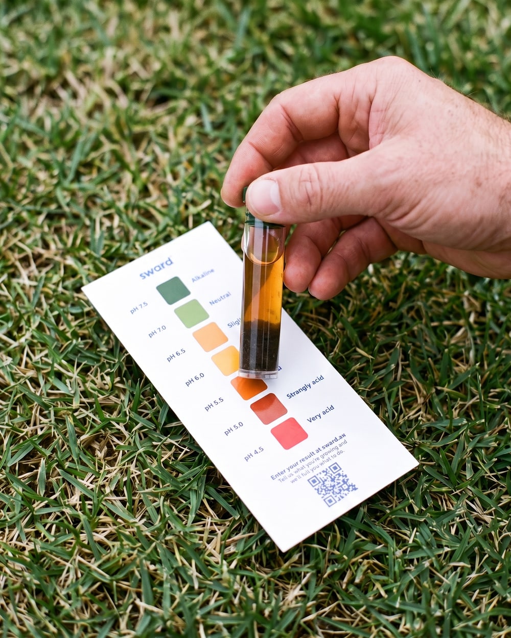 A hand holding a test tube of settled soil against the printed colour chart on a lawn.