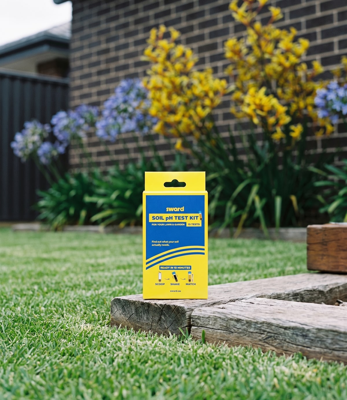 The Sward Soil pH Test Kit box standing on a lawn beside a timber garden edge, with kangaroo paws and agapanthus flowering behind.