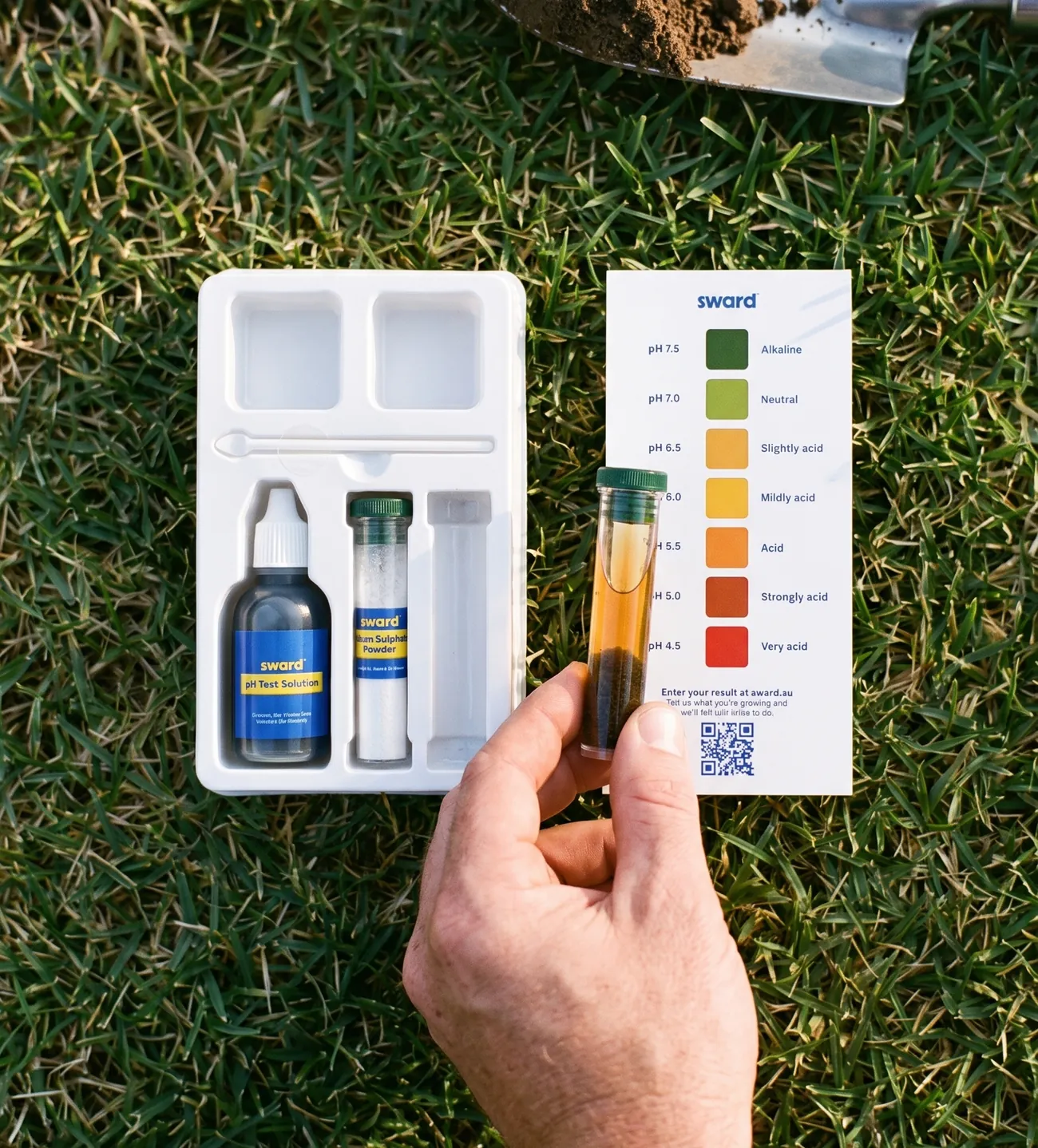 Looking straight down on a lawn: a hand holds a test tube of settled soil against the Soil pH Test Kit colour chart, the open kit beside it and a trowel of soil above.